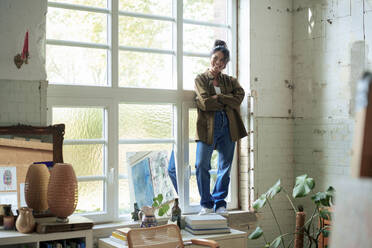 Happy woman with arms crossed standing on window sill at home - JOSEF22749