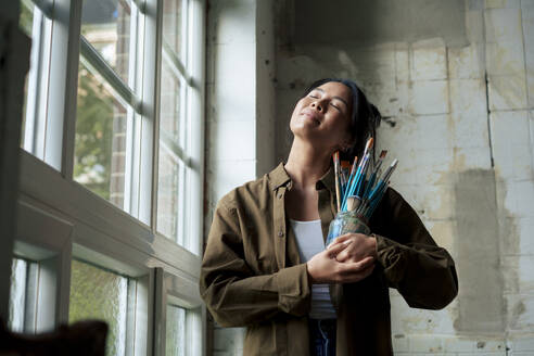 Smiling painter holding paintbrushes with eyes closed standing by window at workshop - JOSEF22754