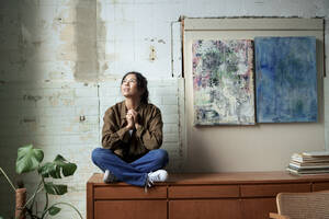 Nachdenklicher Maler, der auf einem Schrank neben Gemälden im Kunstatelier sitzt - JOSEF22757