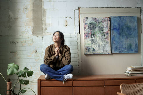 Thoughtful painter sitting on cabinet by paintings at art studio - JOSEF22757