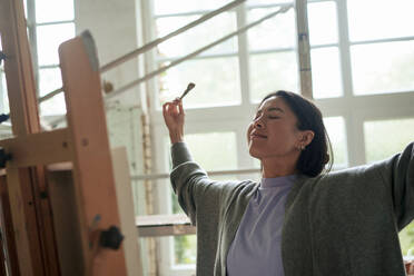 Smiling woman with eyes closed and arms outstretched standing in front of painting at workshop - JOSEF22769