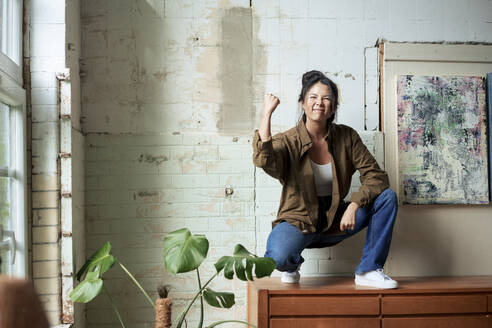 Excited painter showing clenched fist squatting on cabinet by paintings at art studio - JOSEF22770