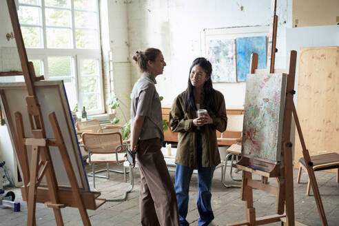 Lächelnde Frauen, die über ein Gemälde auf einer Staffelei im Kunststudio diskutieren - JOSEF22828