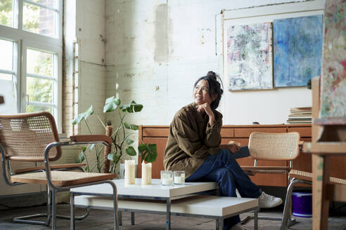 Smiling artist with tablet PC sitting on coffee table day dreaming in workshop - JOSEF22858