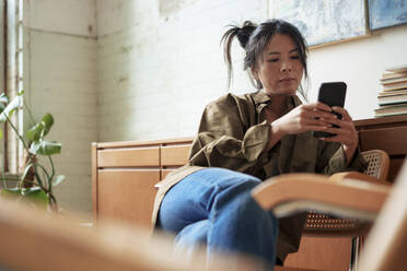 Künstlerin sitzt auf einem Stuhl und benutzt ein Smartphone in einer Werkstatt - JOSEF22859