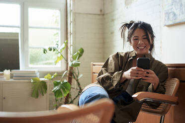 Smiling artist sitting on chair using smart phone in workshop - JOSEF22860