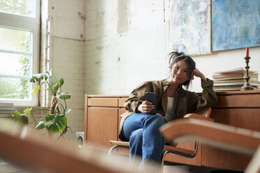 Maler auf einem Stuhl sitzend mit Smartphone in einer Werkstatt - JOSEF22862