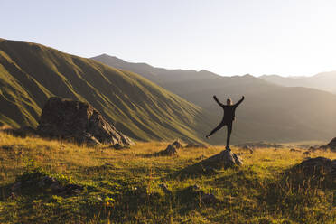 Carefree woman with arms raised standing on rock in front of mountains at sunset - PCLF00942