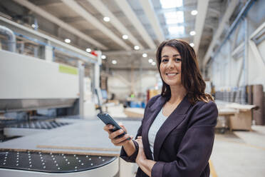 Lächelnde Geschäftsfrau mit Smartphone in der Industrie stehend - JOSEF22884