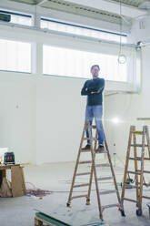 Architect with arms crossed standing on ladder in workshop - JOSEF22935