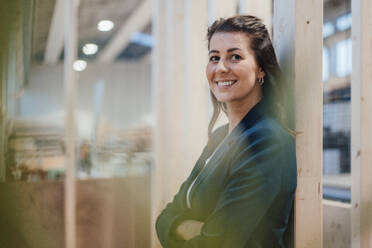 Smiling businesswoman with arms crossed leaning on wooden column - JOSEF22977