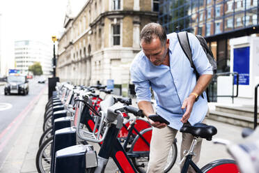 Geschäftsmann, der ein Elektrofahrrad mit Smartphone auf der Straße mietet - WPEF08008