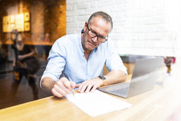 Geschäftsmann schreibt an einem Dokument neben einem Laptop in einem Café - WPEF08036