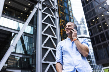 Lächelnder Geschäftsmann, der vor einem Gebäude mit seinem Smartphone telefoniert - WPEF08042