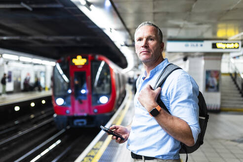 Älterer Geschäftsmann hält Smartphone und wartet auf dem Bahnsteig auf einen Zug - WPEF08074