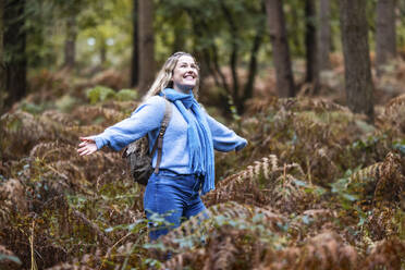 Glückliche junge Frau steht mit ausgestreckten Armen inmitten von Farnpflanzen im Cannock Chase Forest - WPEF08086