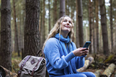 Lächelnde Frau sitzt mit Rucksack und Smartphone im Cannock Chase Forest - WPEF08103