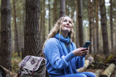 Smiling woman sitting with backpack and smart phone in Cannock chase forest - WPEF08103