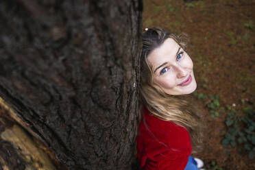 Smiling beautiful woman leaning on tree trunk in forest - WPEF08109