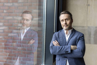 Confident businessman with arms crossed standing in workplace - PESF04156