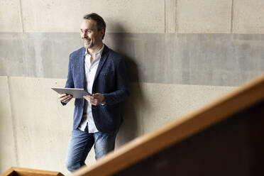 Happy businessman with tablet computer in front of wall - PESF04164