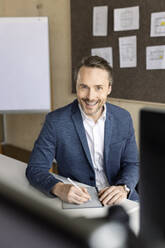 Smiling businessman with tablet PC sitting at desk in workplace - PESF04189