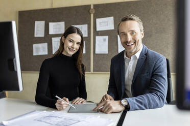 Happy businessman and colleague sitting at desk in workplace - PESF04193