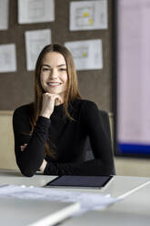 Lächelnder junger Auszubildender mit Hand am Kinn am Schreibtisch am Arbeitsplatz - PESF04200