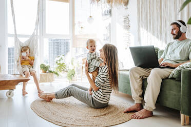 Mutter hält ihren Sohn im Wohnzimmer zu Hause, während der Vater einen Laptop benutzt - WESTF25283