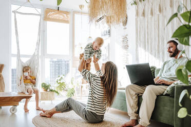 Mother playing with her son in living room at home with father using laptop - WESTF25287