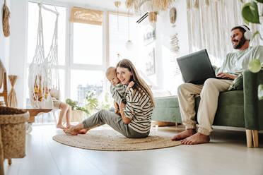 Mother holding with her son in living room at home with father using laptop - WESTF25290