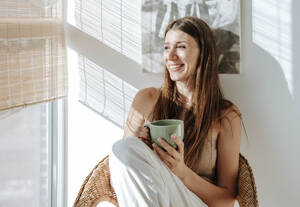 Happy woman with coffee mug relaxing in chair at the window - WESTF25292