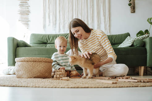 Mutter und Sohn mit Katze spielen im Wohnzimmer zu Hause - WESTF25297