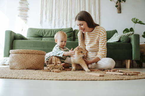 Mutter und Sohn mit Katze spielen im Wohnzimmer zu Hause - WESTF25298