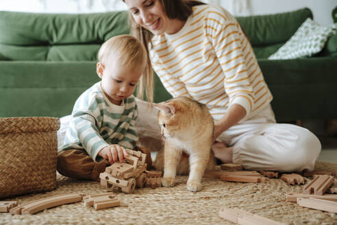 Mutter und Sohn mit Katze spielen im Wohnzimmer zu Hause - WESTF25299