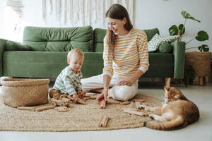 Mutter und Sohn spielen im Wohnzimmer mit einer Holzeisenbahn, beobachtet von einer Katze - WESTF25300