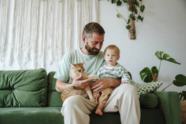 Father and son sitting with cat on the couch in living room at home - WESTF25304