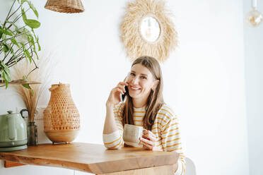 Porträt einer lächelnden Frau mit Kaffeetasse, die zu Hause telefoniert - WESTF25310
