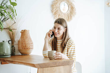 Smiling woman with coffee mug talking on the phone at home - WESTF25311