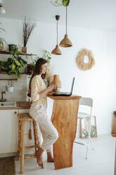 Frau mit Kaffeebecher und Laptop in der Küche zu Hause - WESTF25316