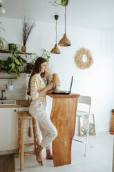 Woman with coffee mug using laptop in kitchen at home - WESTF25316