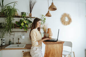 Frau mit Kaffeebecher und Laptop in der Küche zu Hause - WESTF25317