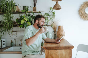 Mann hält Kaffeetasse und benutzt Mobiltelefon zu Hause - WESTF25328