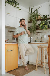 Mann mit Kaffeetasse beim Telefonieren in der Küche zu Hause - WESTF25332