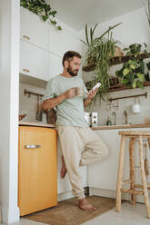 Mann mit Kaffeetasse und Mobiltelefon in der Küche zu Hause - WESTF25333