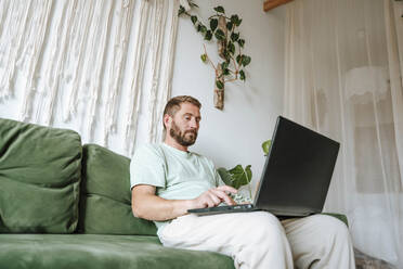 Mann sitzt zu Hause auf der Couch und benutzt einen Laptop - WESTF25341
