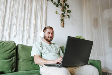 Man with headphones sitting on the couch at home and using laptop - WESTF25342