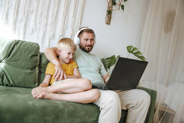 Vater und Sohn schauen auf einen Laptop, während sie zu Hause auf der Couch sitzen - WESTF25345