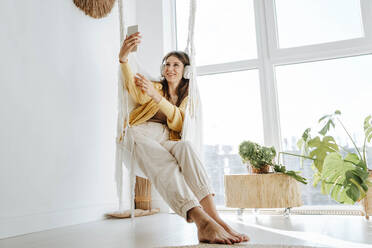 Smiling woman wearing headphones and taking a selfie at home - WESTF25356