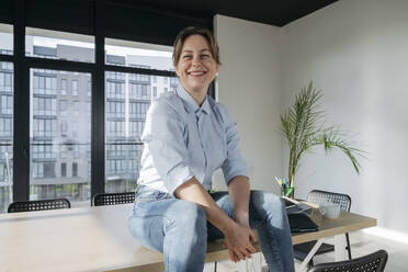 Happy businesswoman sitting on table in office - OSF02349